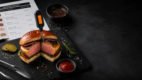 Grilovaný burger s vpíchnutým digitálním teploměrem, tabulkou teplot a tmavým pozadím.