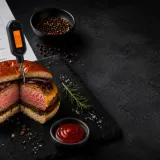 Grilovaný burger s vpíchnutým digitálním teploměrem, tabulkou teplot a tmavým pozadím.