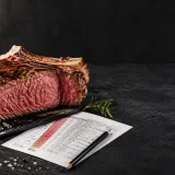 Rozkrojený hovězí steak s růžovým středem na tmavé kamenné desce, do kterého je zapíchnutý digitální teploměr.